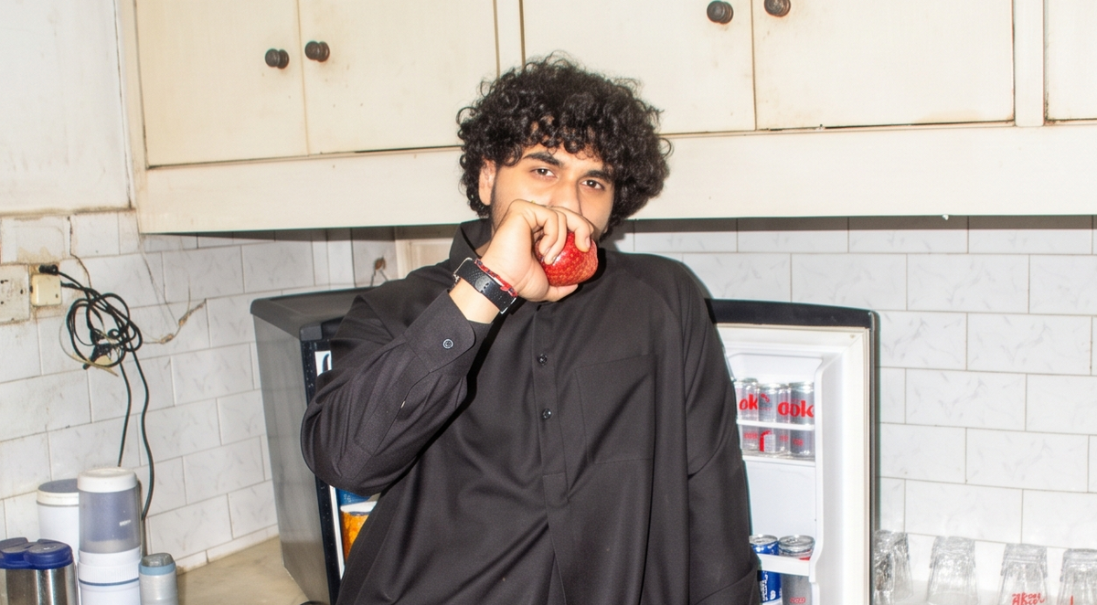 A man with curly black hair wearing a black long-sleeve shirt stands in a kitchen holding a red apple in front of his face. An open refrigerator is visible behind him, along with white cabinets and tiled walls.