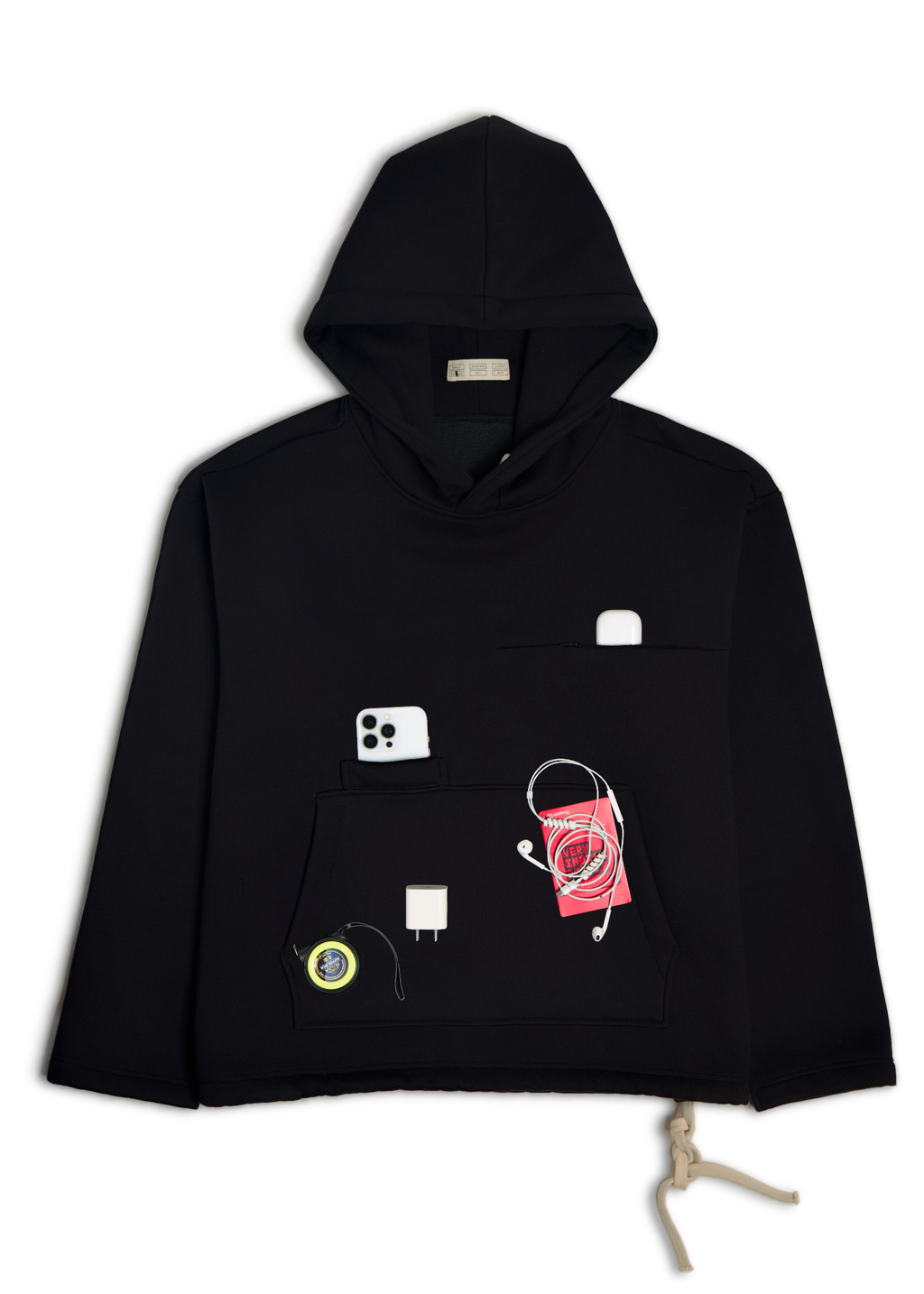 A black cropped hoodie with a kangaroo pocket decorated with various small accessories, including a red patterned pouch, a white USB-C cable, a white rectangular accessory, and a small circular patch, laid flat on a light gray background.