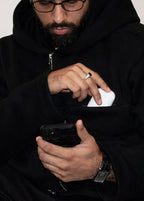Man wearing a black hoodie holding a phone with a white object in his pocket.