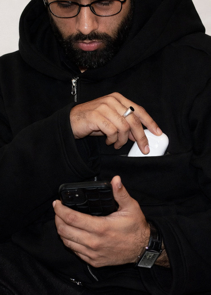 Man wearing a black hoodie holding a phone with a white object in his pocket.
